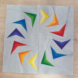 Op de afbeelding: Een patchwork quiltblok met een regenboogontwerp. Het blok is gemaakt van meerdere driehoeken in verschillende kleuren, waaronder rood, oranje, geel, groen, blauw, indigo en violet. De driehoeken zijn in een spiraalvorm gerangschikt.