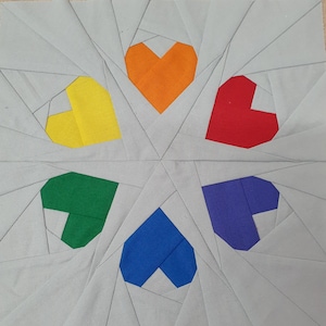 May include: A square fabric block featuring a central design of six colorful hearts in a starburst pattern. The hearts are in shades of yellow, orange, red, green, blue, and purple, set against a gray background with geometric lines.