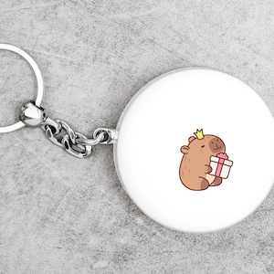 May include: A white keychain with a silver key ring and a round charm featuring a cartoon animal wearing a crown and holding a gift box.