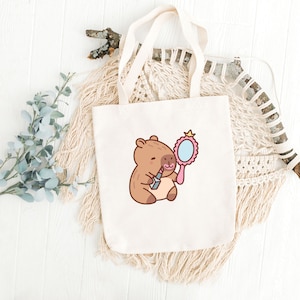 May include: A white tote bag with a cartoon illustration of a capybara holding a lipstick and a pink hand mirror. The capybara is brown with a pink nose and a pink blush on its cheeks.