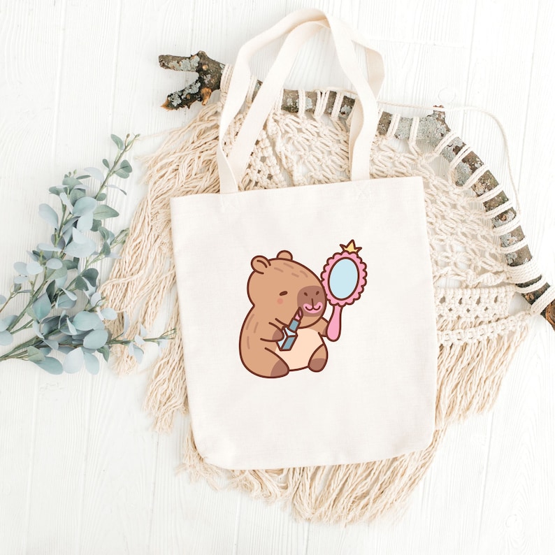 May include: A white tote bag with a cartoon illustration of a capybara holding a lipstick and a pink hand mirror. The capybara is brown with a pink nose and a pink blush on its cheeks.