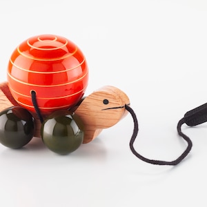 Puede incluir: Una tortuga de juguete de madera con una bola rayada roja y amarilla en la espalda. La tortuga tiene tres bolas verdes para las patas y una cuerda negra atada a la cabeza.
