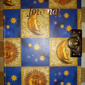 Puede incluir: Un diario azul y dorado con un candado y una llave. La portada presenta un patrón repetitivo de soles y lunas con estrellas. La palabra "Journal" está escrita en letras doradas en la portada.