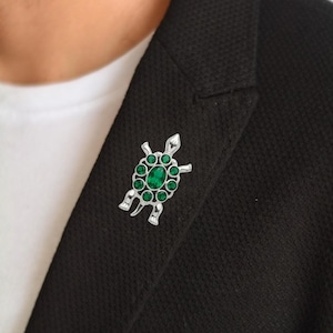 May include: A silver-toned turtle brooch featuring a large central oval green gemstone surrounded by eight smaller round green stones set into the shell. The metal frame is polished and smooth, forming the head, four legs, and a small tail.