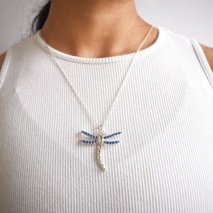 Puede incluir: Un collar de plata con un colgante de libélula. La libélula está adornada con cristales azules.