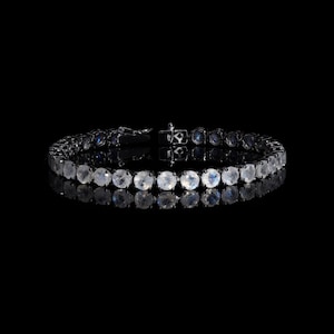 May include: A silver bracelet with a row of round, white moonstone gems set in a black metal band. The bracelet is reflected in a black surface.