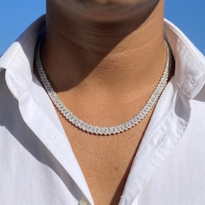 May include: A silver-colored, diamond-studded necklace. The necklace is a cuban link style, with multiple rows of small, sparkling stones. The necklace is worn against a white collared shirt, with a blue sky in the background.