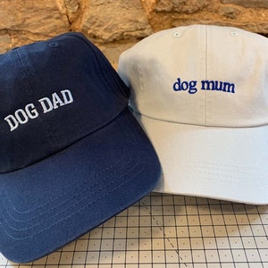 May include: Two baseball caps, one navy blue with white embroidery that says "DOG DAD" and one beige with blue embroidery that says "dog mum".