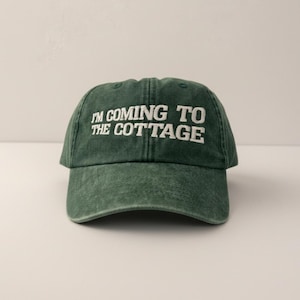 May include: A dark green baseball cap with the embroidered text "I'M COMING TO THE COTTAGE" in white. The cap has a curved brim and a slightly faded appearance. A classic, casual hat, perfect for a relaxed, cottage-themed aesthetic.