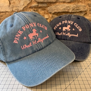 May include: Two blue denim baseball caps with pink embroidered logos. The logos say "Pink Pony Club West Hollywood" and feature a pink silhouette of a carousel horse.