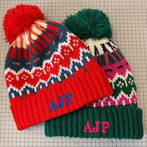 May include: Two knitted winter hats with pom-poms. One hat is red with blue and white patterns and blue embroidered initials. The other is green with pink and white patterns and pink embroidered initials. Both hats have a ribbed knit brim.