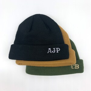 May include: Three knitted beanies stacked on top of each other. The top beanie is black with white embroidered letters that say "AJp". The middle beanie is brown and the bottom beanie is green with gold embroidered letters that say "CB".