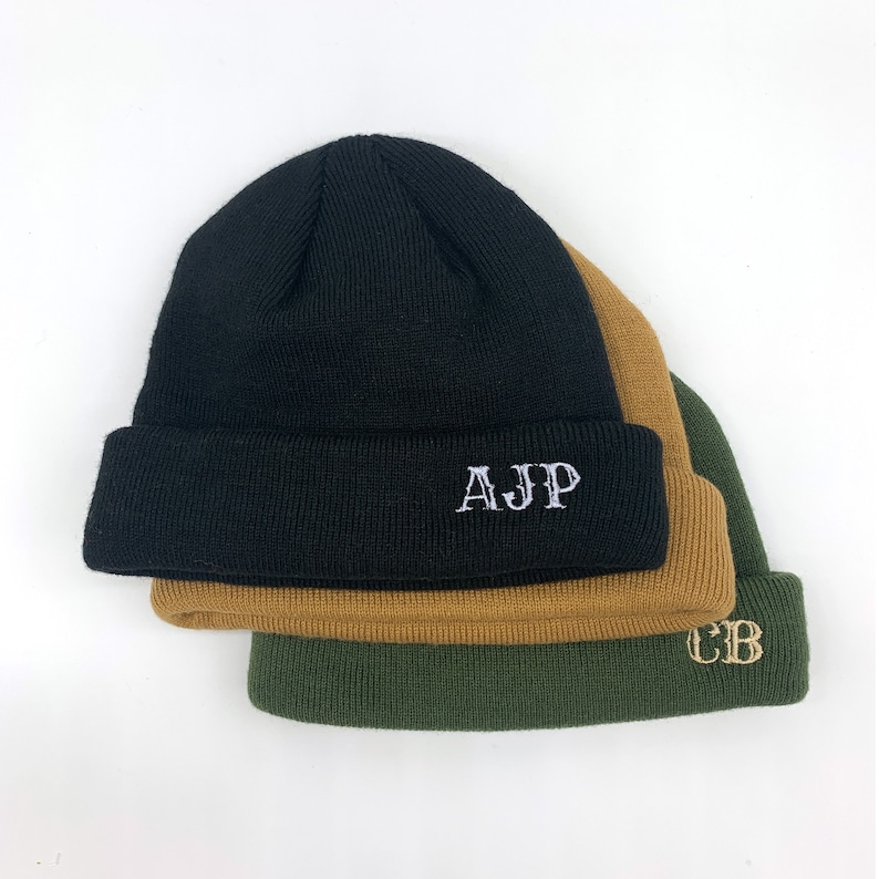 May include: Three knitted beanies stacked on top of each other. The top beanie is black with white embroidered letters that say "AJp". The middle beanie is brown and the bottom beanie is green with gold embroidered letters that say "CB".
