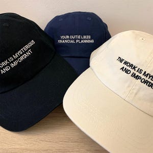 May include: Three baseball caps with different embroidered text. The first cap is black with white text that reads "Work is mysterious and important". The second cap is blue with white text that reads "Your outie likes financial planning". The third cap is beige with black text that reads "The work is mysterious and important".