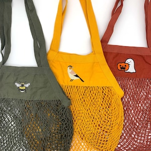 May include: Three reusable shopping bags with different embroidered designs. The bag on the left is olive green with a bee design. The middle bag is yellow with a bird design. The bag on the right is red with a pumpkin and ghost design.