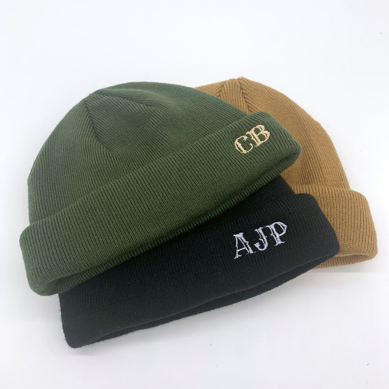 May include: Three knitted beanies in different colors, green, black, and brown. The beanies have embroidered initials on them, "CB", "AJ", and "P".