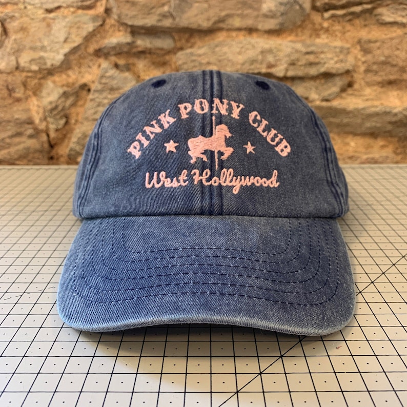 May include: A blue denim baseball cap with the text "Pink Pony Club West Hollywood" embroidered in pink. A pink silhouette of a carousel horse is in the center of the text.