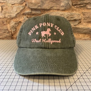 May include: A dark green baseball cap with the text "Pink Pony Club West Hollywood" embroidered in pink. A pink silhouette of a carousel horse is also embroidered on the cap.