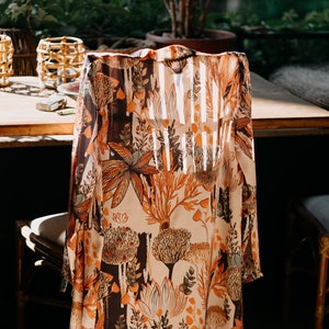 Puede incluir: Prenda larga y fluida con estampado botánico de árboles y follaje en tonos naranja, marrón y crema. La prenda está drapeada sobre una silla, mostrando su diseño y tejido.