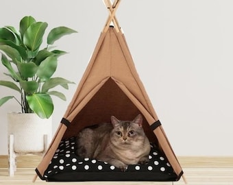 May include: A tan fabric pet teepee tent with wooden support poles at the top. Inside the tent, a grey cat rests on a black cushion featuring a white polka dot pattern. The structure has an open front entrance and black fabric ties on the sides.