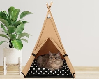 May include: A tan fabric pet teepee tent with wooden support poles at the top, featuring an open front entrance. Inside the tent, a grey cat rests on a black cushion with a white polka dot pattern. The structure is triangular in shape.