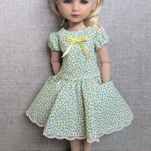 Könnte beinhalten: Eine Puppe in einem grünen Blumenkleid mit gelber Schleife und weißen Schuhen. Das Kleid hat kurze Ärmel und einen ausgestellten Rock. Die Puppe hat blondes Haar und ein gelbes Stirnband. Das Kleid ist mit weißer Spitze verziert.