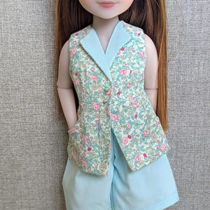 Puede incluir: Una muñeca vestida con un chaleco con estampado floral con cuello azul claro y pantalones cortos a juego. El chaleco tiene rosas rosas y hojas verdes. La muñeca tiene pelo largo color castaño rojizo y zapatos blancos.