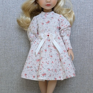 May include: A doll with blonde hair and a floral print dress. The dress has a white lace collar and a white bow with a red accent. The doll is wearing white shoes.