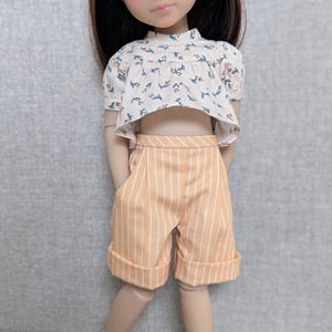 Op de afbeelding: Een pop met bruin haar draagt een wit shirt met korte mouwen en een klein bloemenpatroon, gecombineerd met perzikkleurige shorts met witte verticale strepen en omgeslagen zomen. De pop draagt ook witte schoenen met gespen en kleine details.