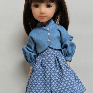 Peut inclure: Poupée vêtue d'une veste en jean bleue avec des boutons roses et d'une jupe assortie à motif floral blanc. La poupée a les cheveux bruns et porte des collants beiges.