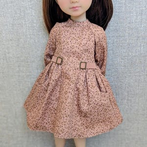 Könnte beinhalten: Puppe mit braunen Haaren und grünen Augen, die ein rosa geblümtes Kleid mit passendem Gürtel und Schuhen trägt. Das Kleid hat lange Ärmel und einen hohen Kragen. Die Puppe steht.