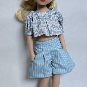 May include: A doll with blonde hair wearing a short-sleeved crop top featuring a blue and pink floral pattern, paired with light blue striped shorts that have front pockets and small decorative buttons, and white buckled shoes.