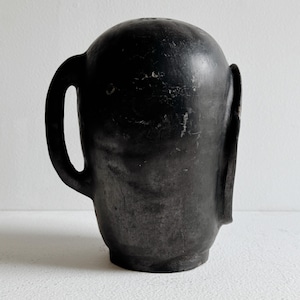 May include: A black ceramic mug with a rounded top and a handle on the left side. The mug has a textured surface and a small spout on the right side. The mug is sitting on a white surface.