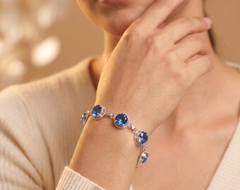Puede incluir: Una pulsera de tono plateado que presenta una serie de piedras preciosas azules redondas y facetadas, engastadas en biseles metálicos individuales. Las piedras están unidas por pequeños anillos de metal plateado, creando un diseño flexible.