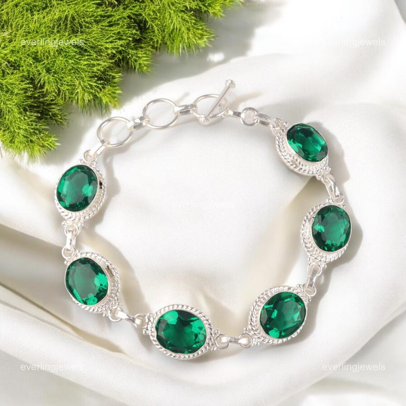 May include: A silver bracelet featuring oval-cut, vibrant green gemstones. Each gem is set in a decorative silver frame, connected by delicate links. The bracelet has a toggle clasp and is displayed on a white surface.