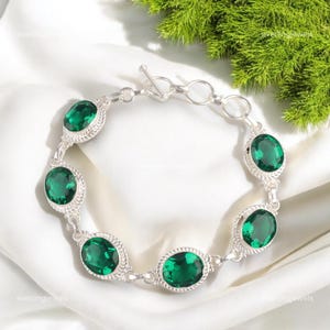 May include: A silver bracelet featuring oval-cut, emerald-green gemstones. Each gem is set in a decorative silver frame, connected by delicate links. The bracelet is displayed on a white surface with green foliage in the background.