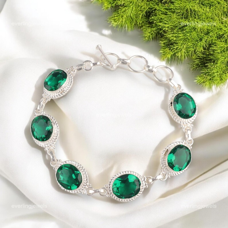 May include: A silver bracelet featuring oval-cut, emerald-green gemstones. Each gem is set in a decorative silver frame, connected by delicate links. The bracelet is displayed on a white surface with green foliage in the background.