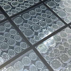 May include: Multiple sheets of clear adhesive nail tabs for applying false nails. The tabs are arranged in rows on a black background.