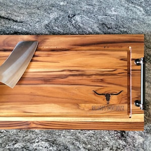 May include: A large wooden cutting board with a black metal handle on each end. The board has a groove around the perimeter and a smaller groove in the center. A silver cleaver with a black and red handle is resting on the board. The board is engraved with a bull skull and the text 'Richard's Smokehouse'.