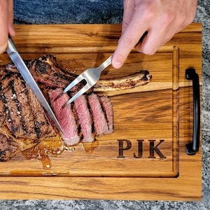 May include: A large, grilled ribeye steak being sliced on a wooden cutting board. The steak is seasoned with salt and has grill marks. The board has black handles and the initials "PJK" carved into it.