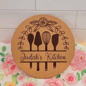 May include: A round cork trivet with a floral design and four kitchen utensils, including a spoon, spatula, whisk, and turner. The text "Judah's Kitchen" is engraved below the utensils.