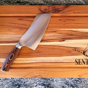 May include: A rectangular wooden cutting board with a built-in juice groove and dark metal handles. A large chef's knife with a patterned wooden handle rests on the board. The board is engraved with a flying goose design and the word "SENTER".