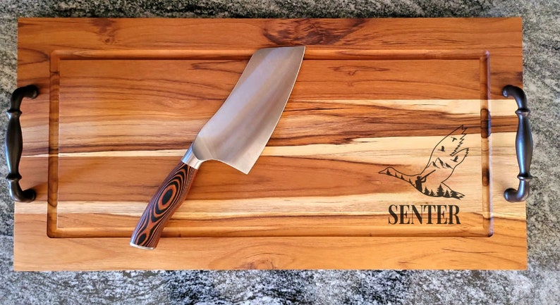 May include: A rectangular wooden cutting board with a built-in juice groove and dark metal handles. A large chef's knife with a patterned wooden handle rests on the board. The board is engraved with a flying goose design and the word "SENTER".