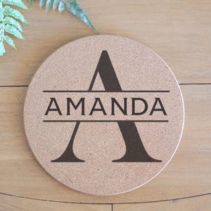 May include: Round cork coaster with the name "AMANDA" printed in black, bisected by a large letter "A". The coaster is light brown and sits on a wooden surface. A fern leaf is in the background.