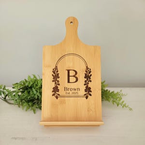 May include: A personalized bamboo cutting board with a floral arch design, the initial "B" and the text "Brown Est. 2025" engraved on the surface.