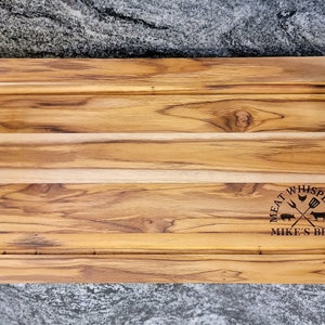 May include: A rectangular wooden cutting board with a juice groove and black metal handles. The board has the text "MEAT WHISPERER" and "MIKE'S BBQ" etched into the wood. The wood grain is visible.