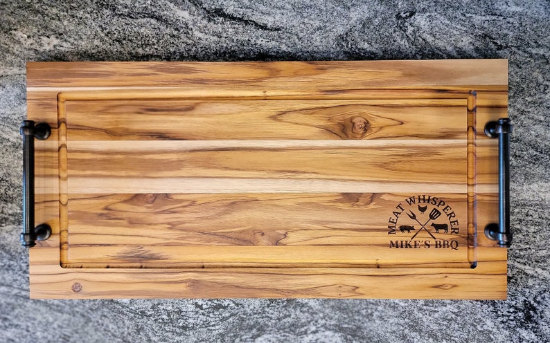 May include: A rectangular wooden cutting board with a juice groove and black metal handles. The board has the text "MEAT WHISPERER" and "MIKE'S BBQ" etched into the wood. The wood grain is visible.