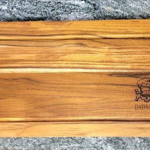 May include: A rectangular wooden cutting board with a carved groove and black handles. The board features a skull and crossbones graphic wearing a chef's hat, with the text "DAD'S BBQ" below.
