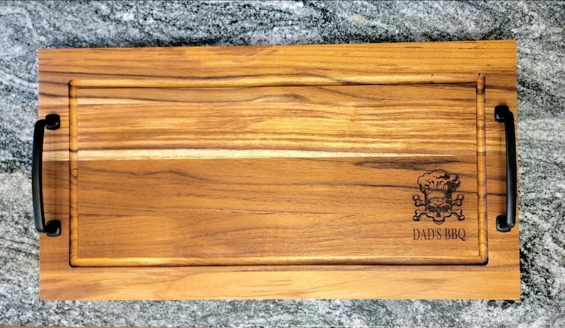 May include: A rectangular wooden cutting board with a carved groove and black handles. The board features a skull and crossbones graphic wearing a chef's hat, with the text "DAD'S BBQ" below.