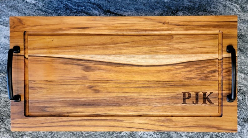 May include: A rectangular wooden serving board with a light brown finish and dark wood grain. It features a carved groove, black handles, and the initials "PJK" engraved on the surface. The board is designed for serving food.